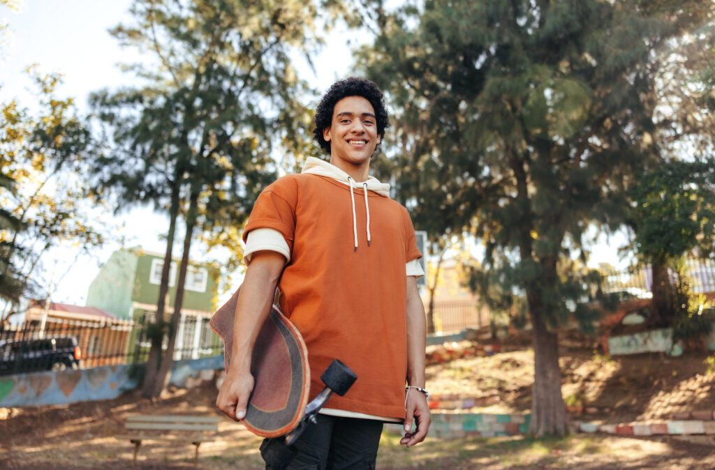 Carefree skateboarder holding his skateboard in an urban park. Cheerful teenage boy smiling at the camera while standing outdoors. Sporty teenager wearing casual clothing during the day.